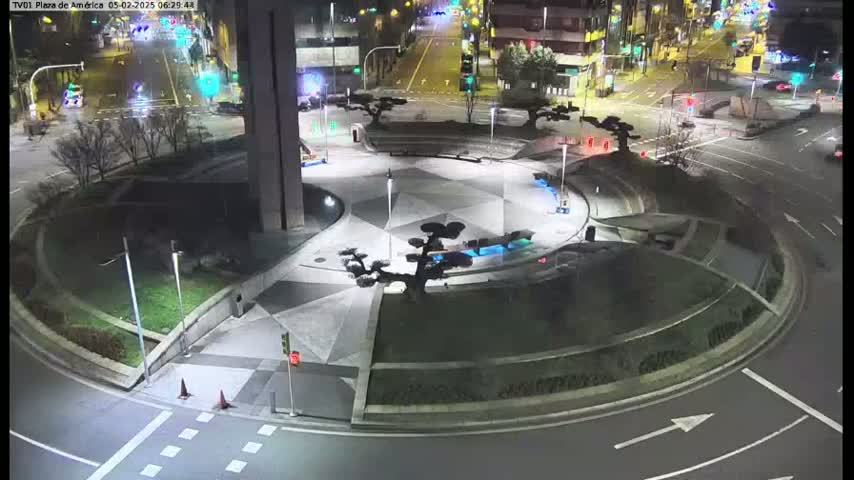 A nighttime aerial view of a circular plaza with landscaping and artificial trees, situated at a roadway intersection with sparse traffic.