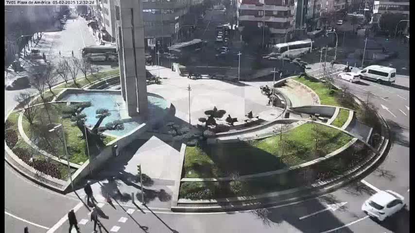 A sunny day shows a city plaza with a circular landscaped area featuring a fountain, trees, and benches, surrounded by a roadway with some pedestrian and vehicle traffic.