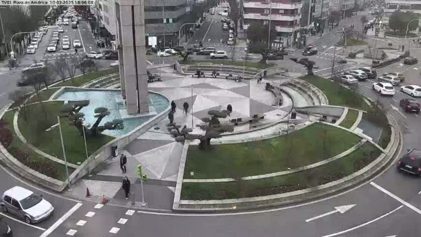 A roundabout with a modern, landscaped center is surrounded by city streets with cars driving on an overcast day.