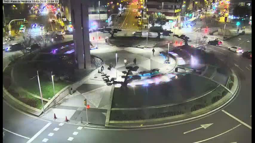 A nighttime aerial view of a circular plaza with a few pedestrians and cars driving around it.
