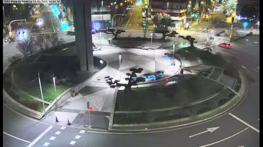 A nighttime aerial view shows a paved roundabout with landscaping, streetlights, and a few cars driving around it.