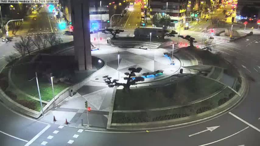 An illuminated plaza at night with geometric landscaping, small trees, and surrounding roadways.
