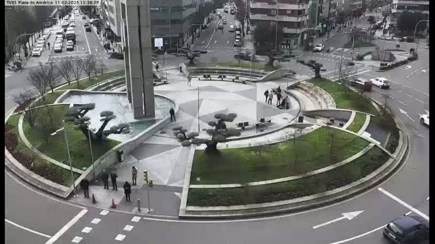 A circular plaza with landscaping features, including water features and sculpted trees, is situated in the center of a roundabout with cars driving around it on a mostly overcast day.