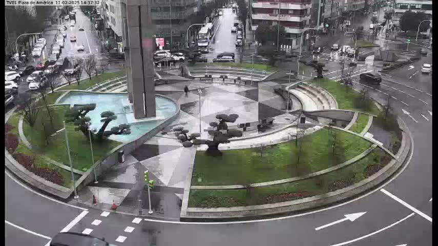 A wet, landscaped traffic circle in a city, featuring a central fountain and geometrically designed gardens, is surrounded by streets with cars and buildings.