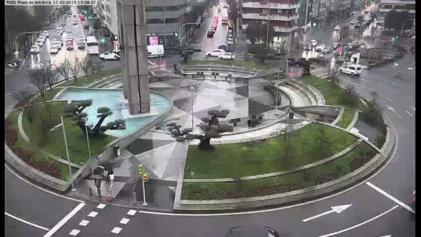 A rain-slicked roundabout features a central plaza with geometric landscaping, a reflecting pool, and several sculpted trees.