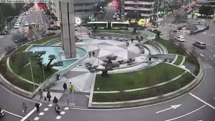 A circular plaza with a geometric design, featuring landscaping, a fountain, and several people walking, is surrounded by roadways with cars, under an overcast sky.