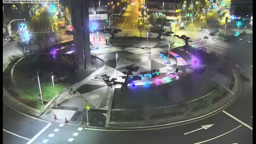 A nighttime aerial view of a geometrically patterned roundabout with landscaping and streetlights, surrounded by roadways and buildings.