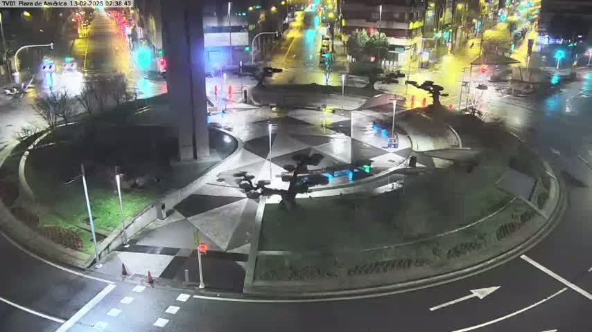 A wet, nighttime aerial view of a geometrically designed roundabout with sparse traffic and landscaping.