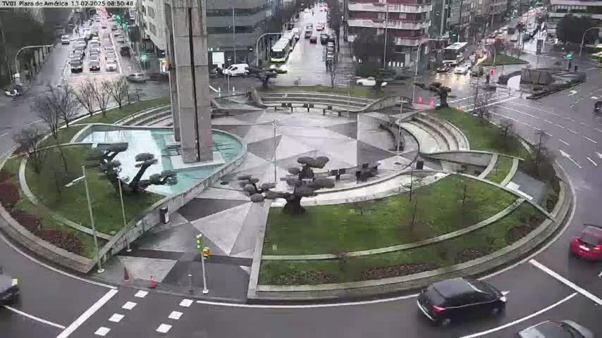 A circular plaza with geometric landscaping and several small trees sits in the center of a roundabout, with cars moving around it in wet conditions.