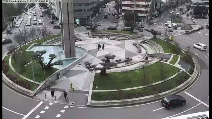 An aerial view of a circular plaza with a fountain and landscaping, surrounded by a roadway with cars, on an overcast day.
