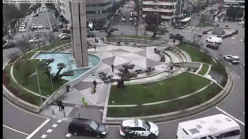 A circular plaza with a geometric pattern, water features, and sculpted trees is surrounded by roads with cars and a bus, under an overcast sky.