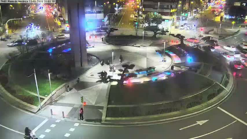 A nighttime aerial view of a roundabout with illuminated water features, surrounded by streets with cars and pedestrians, under partly cloudy conditions.