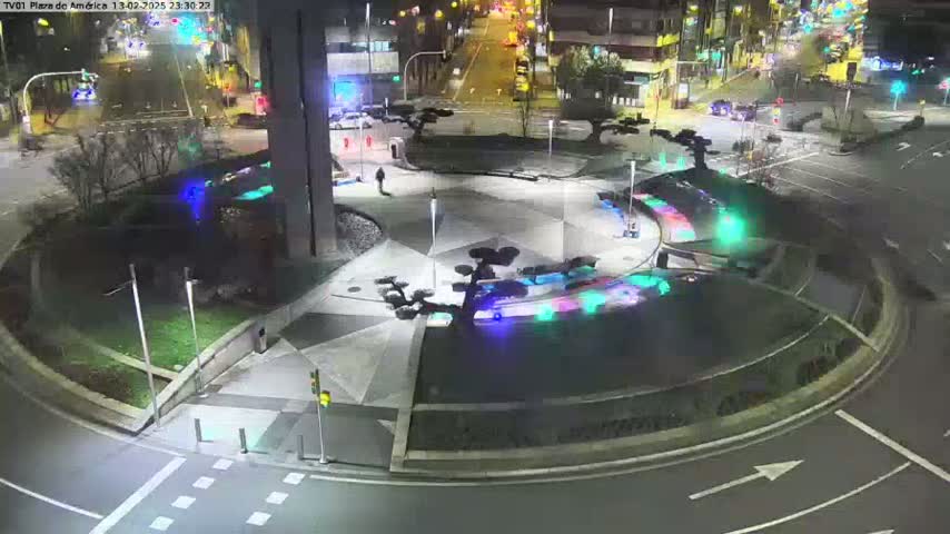 A nighttime aerial view of a roundabout with colorful lights illuminating a central plaza, surrounded by roadways and sparsely-lit buildings.