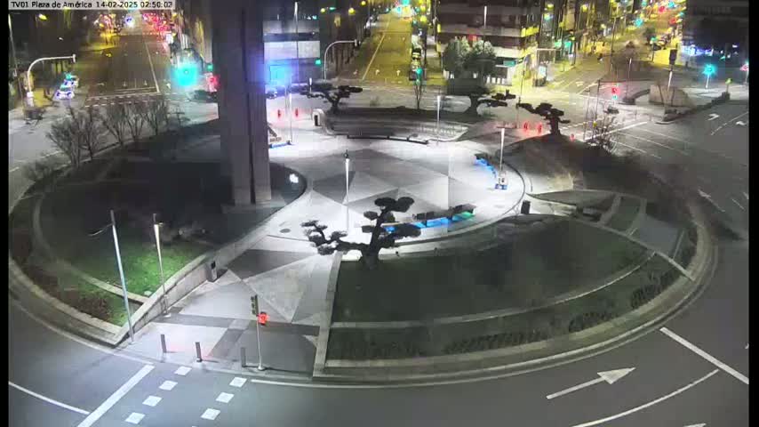 A nighttime aerial view shows a circular plaza with landscaped areas and small trees, situated at a roadway intersection.