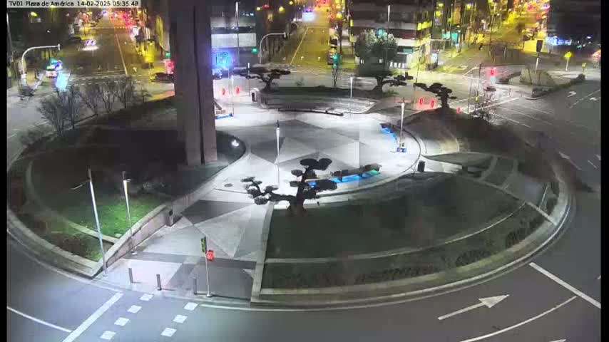 A nighttime aerial view shows a circular plaza with geometric landscaping, small trees, and surrounding roadways with traffic.