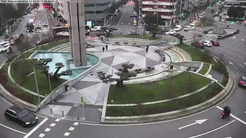 A circular plaza with landscaping, a fountain, and several small trees is situated at the intersection of city streets on an overcast day.