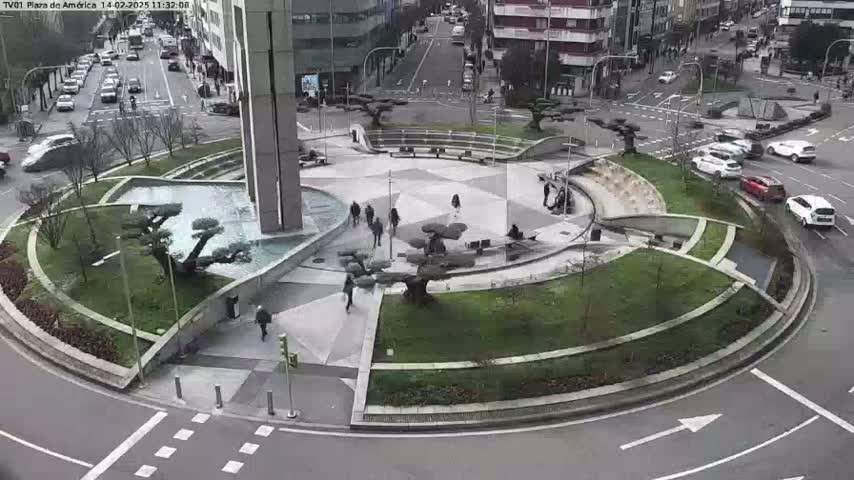 A circular plaza with a fountain and landscaping is surrounded by a roadway on a partly cloudy day, with several people walking and cars driving nearby.