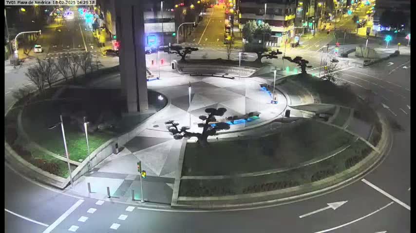 An illuminated nighttime roundabout with landscaped features, including several small trees, is surrounded by roads with minimal traffic.