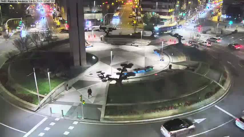 A nighttime view of a roundabout with cars driving around it,  surrounded by landscaped areas and a building, under illuminated streetlights.