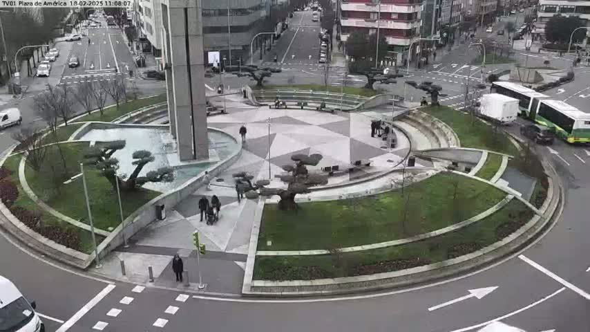 A roundabout in a city with a geometrically designed center featuring landscaping and a fountain, surrounded by streets with cars and buses, on a cloudy day.