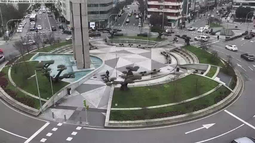An aerial view of a circular plaza with a fountain and landscaping, surrounded by roads with cars, on an overcast day.