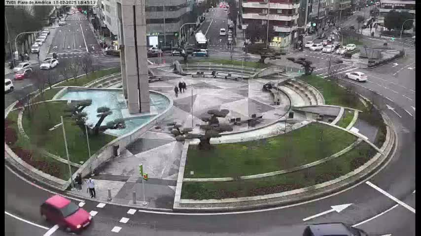 A wet roundabout in a city center features a central plaza with fountains and landscaping, surrounded by roads with cars.