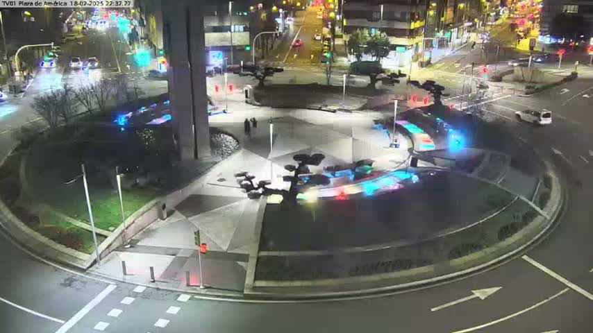 A nighttime aerial view of a roundabout with illuminated landscaping features, surrounded by roads with sparse traffic.