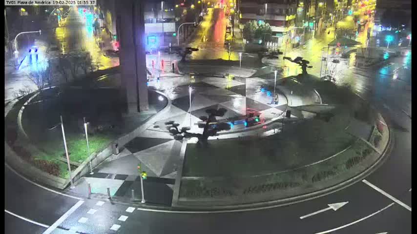 A wet, illuminated roundabout at night, featuring geometric landscaping and several small trees, is surrounded by roadways.