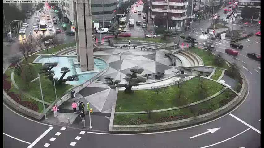 A landscaped roundabout with a central fountain and several trees is surrounded by a roadway with cars and pedestrians under overcast skies.