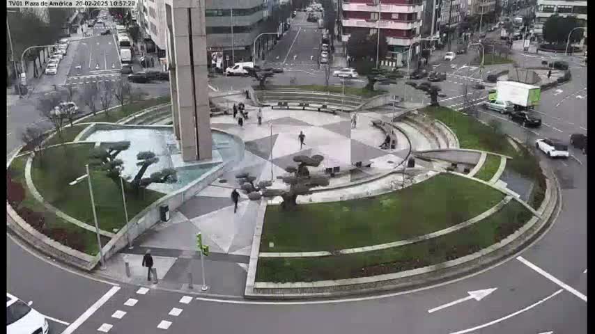 An aerial view shows a roundabout with a landscaped center featuring trees and geometric patterns, surrounded by a city street with cars and pedestrians on an overcast day.