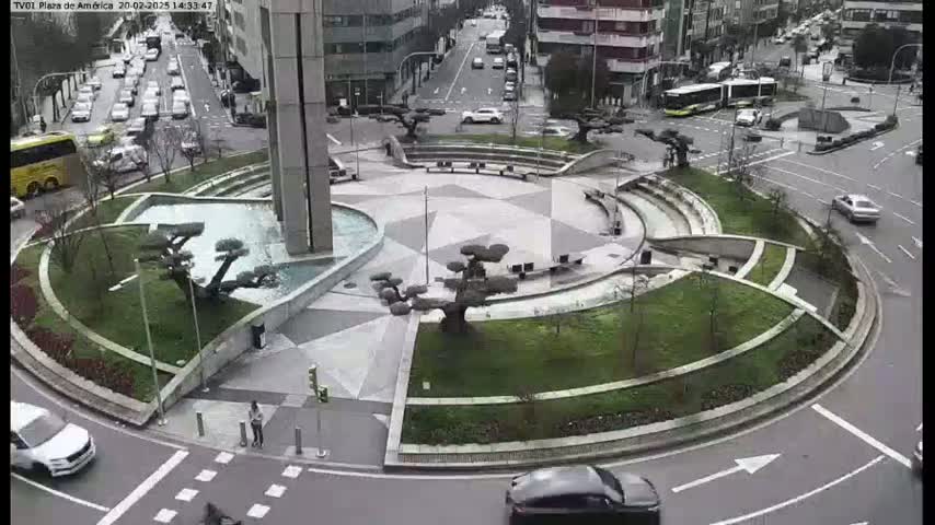A circular plaza with a central fountain and landscaping is surrounded by roadways with cars and buses, under an overcast sky.
