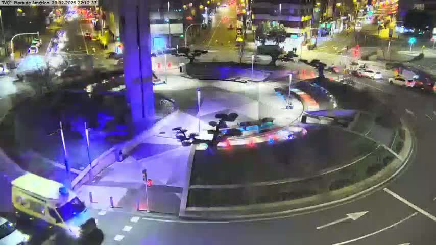 A nighttime aerial view of a roundabout with several vehicles, including an ambulance, and illuminated landscaping features.