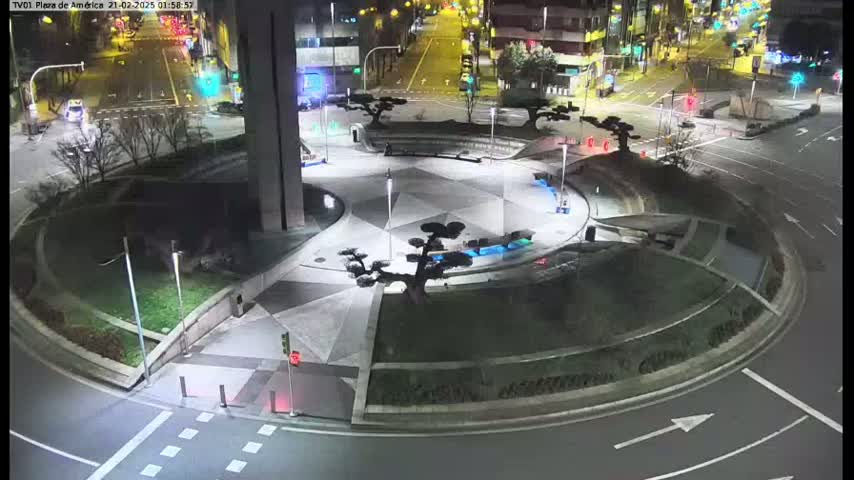 A nighttime aerial view shows a circular plaza with landscaped areas and decorative trees, situated at the intersection of roads with sparse traffic.