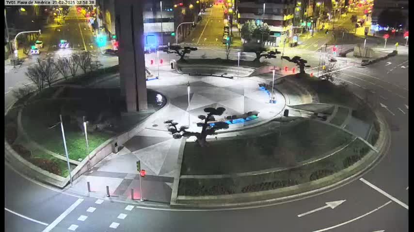 A nighttime aerial view shows a circular plaza with landscaping and several trees, situated in the middle of a roadway intersection.