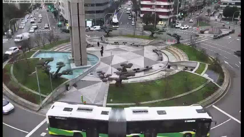 A city roundabout with a central plaza featuring a water feature and landscaping, a bus approaching the roundabout, and light traffic on surrounding roads, under overcast skies.