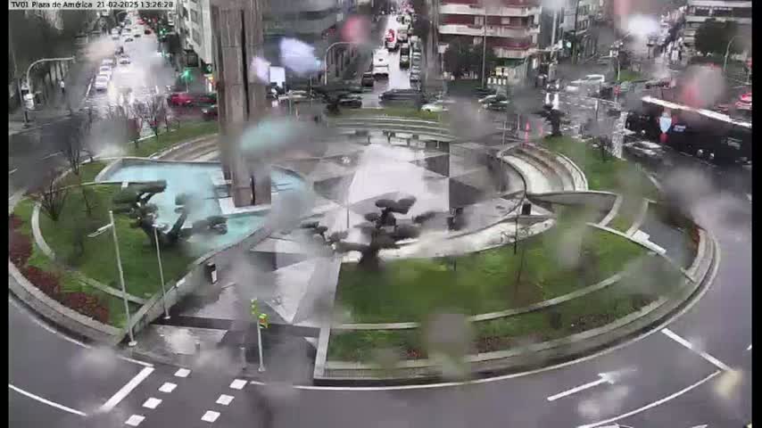 A rain-soaked roundabout features a central fountain and landscaping, surrounded by roadways with cars driving through it.