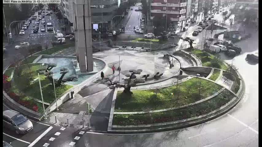 A wet, urban plaza with a circular design featuring landscaping, a fountain, and several people walking around, is seen from an elevated view.