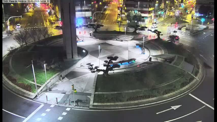 A nighttime view of a roundabout with landscaped islands,  trees, and low lighting, surrounded by streets with cars.