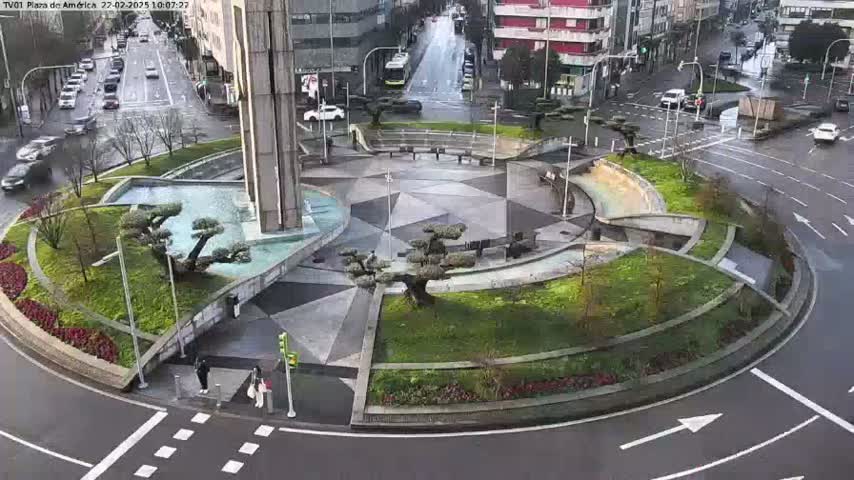 A circular landscaped traffic island, featuring geometric paving, small trees, and a fountain, sits in the center of a wet roadway surrounded by city buildings and cars.