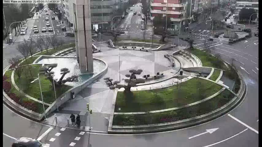 A roundabout with a central plaza featuring a fountain and sculpted trees, surrounded by landscaped areas and buildings, on a wet day.