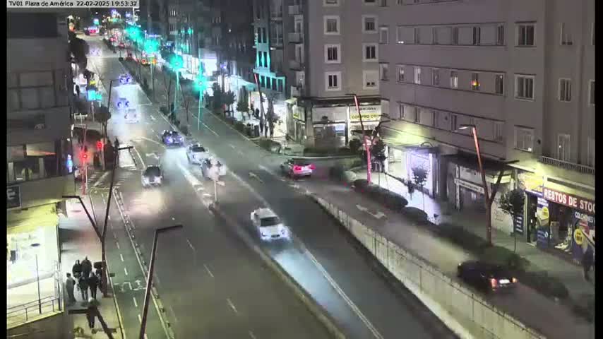 A nighttime view of a city street with several cars driving, buildings lining both sides of the road, and a few pedestrians near the street's edge.