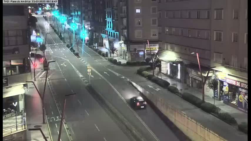 A nighttime, high-angle view of a mostly empty city street with several buildings lining both sides, and a single car driving down the road.