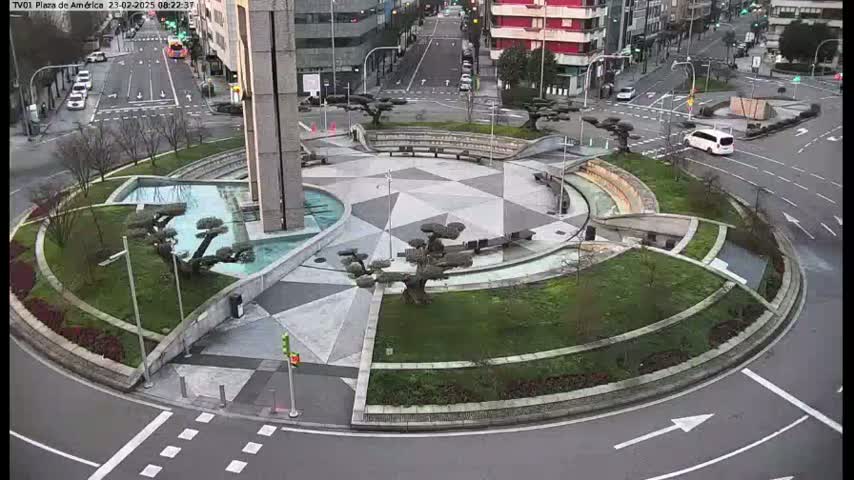 A paved roundabout with landscaped islands featuring water features and small trees is surrounded by a roadway with a few cars.