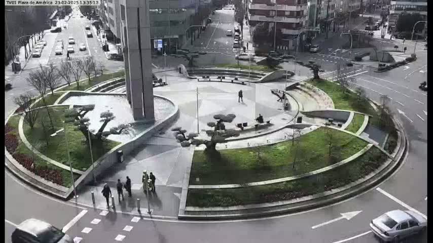 A landscaped traffic circle with a central plaza and several small trees, surrounded by roads with cars and buildings in the background on a partly sunny day.