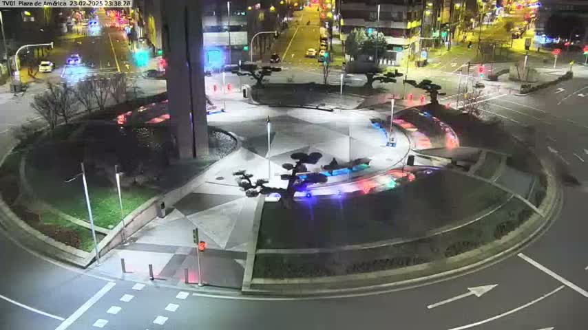 A nighttime aerial view of a roundabout with landscaped areas and illuminated features, surrounded by roads and buildings.