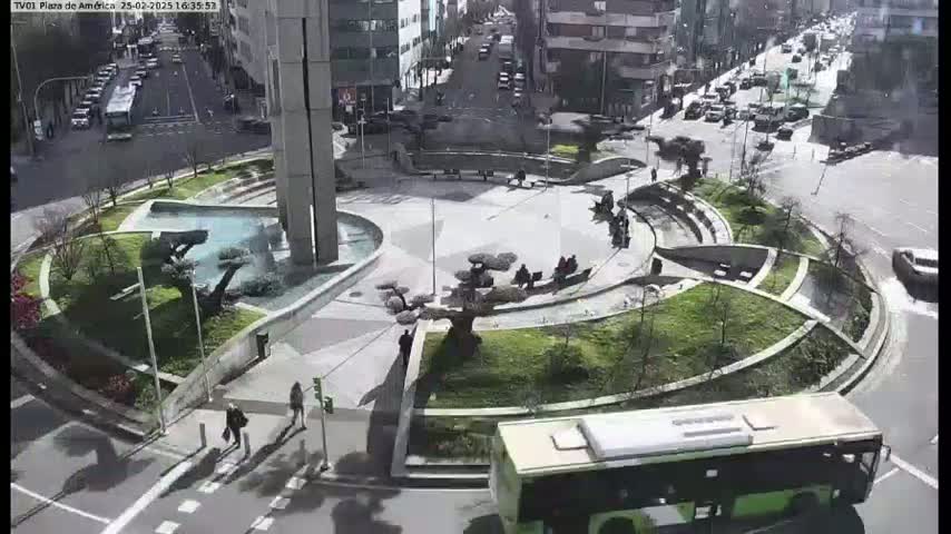 A plaza with a fountain and landscaping is surrounded by buildings and a street with cars, under a sunny sky.