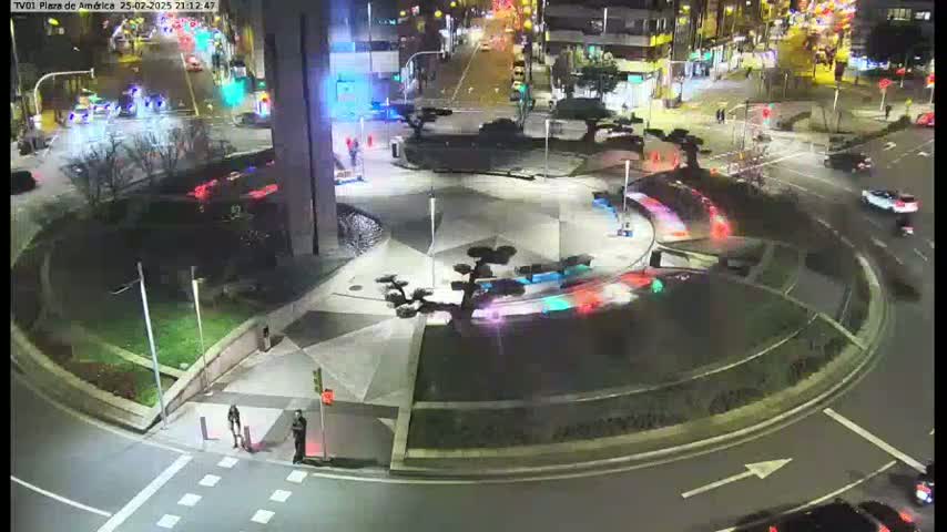 A nighttime aerial view of a roundabout with several cars and pedestrians, surrounded by landscaping and buildings.
