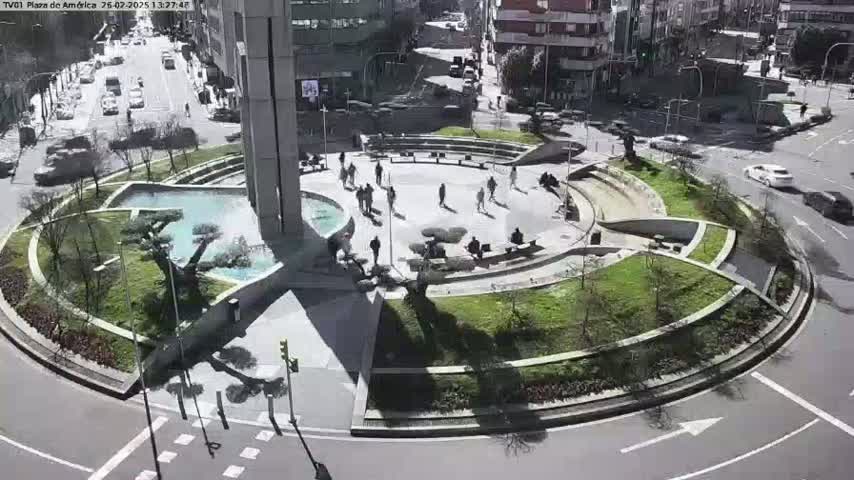 A sunny day shows a circular plaza with landscaping, a fountain, and several people walking around, situated in the middle of a roadway with cars driving around it.