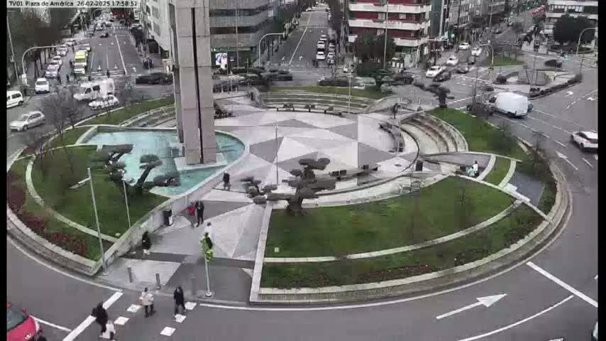 A circular plaza with a geometric pattern, fountains, and landscaping is situated in the center of a traffic circle, with pedestrians and vehicles visible on the surrounding roads under an overcast sky.