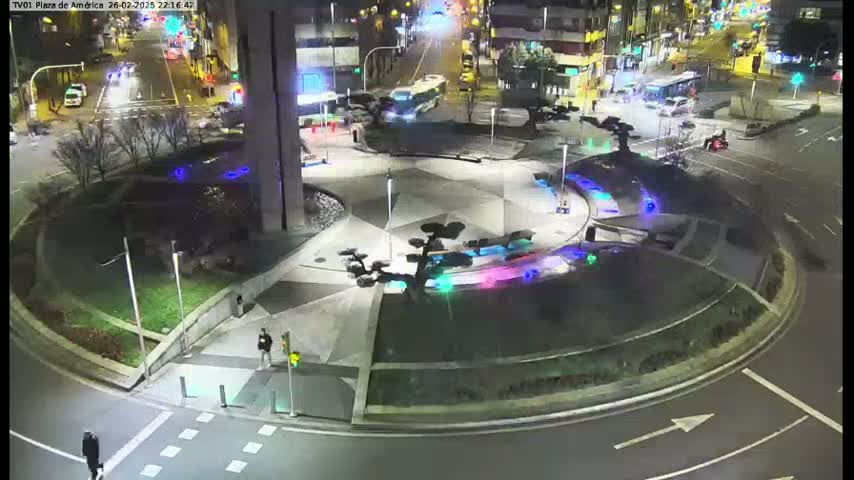 An illuminated roundabout at night, surrounded by roadways and buildings, with a few pedestrians visible.